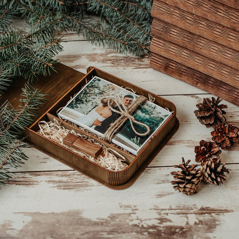 Eine rustikale Hochzeitsfotobox mit USB 3.0, die das Foto eines fröhlichen Paares zeigt, ordentlich mit Bindfaden zusammengebunden, inmitten frischer Tannenzweige, Holzdekor und Tannenzapfen auf einer verwitterten Holzoberfläche.