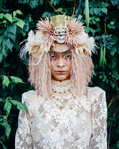 Woman wearing high necked lace vintage dress and crispy rose gold wig, with a skull headdress stares fiercely into camera