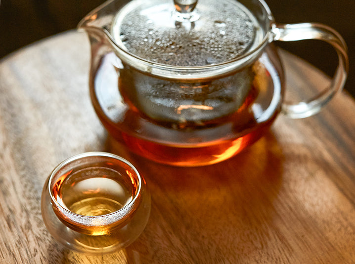 hot brewed hojicha tea in a double walled glass teacup next to a glass infuser teapot