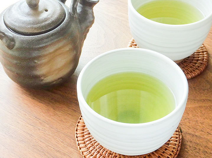 two brewed japanese green tea in white glazed ceramic tea cups on a woven bamboo coasters and kyusu teapot beside