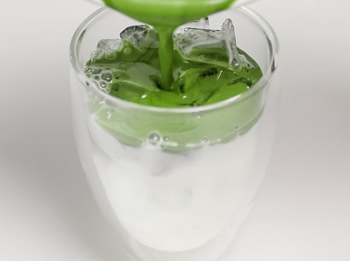 pouring whisked matcha into the double walled glass cup filled with ice and milk to make a cold matcha tea latte beverage