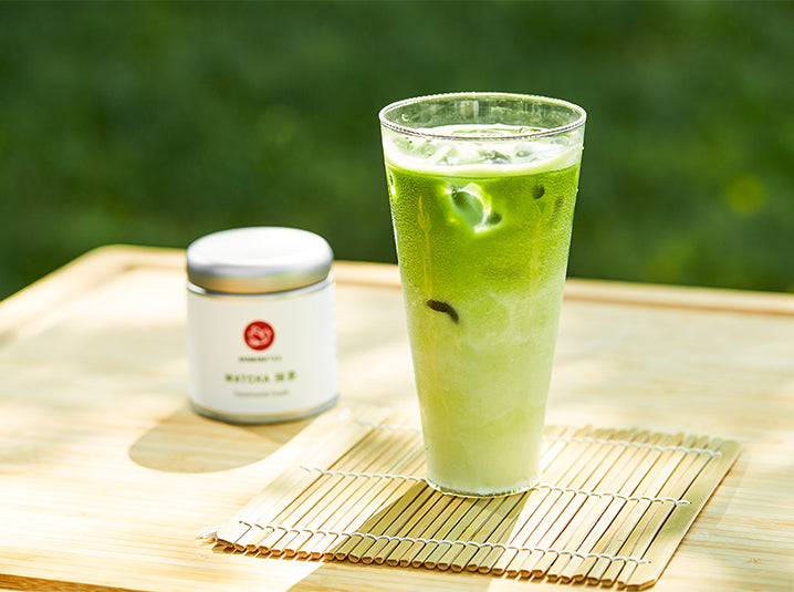 iced matcha green tea latte in a slim tall glass cup on top of a bamboo coaster with senbird tea's matcha otome tin outdoors a staple in the global matcha market