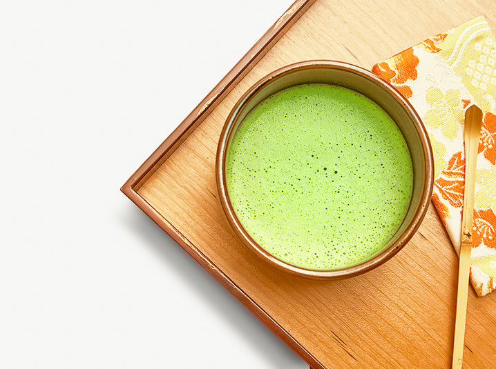 whisked matcha green tea in a brown ceramic bowl with floral designed tenugui and bamboo scoop on top of a wooden tray