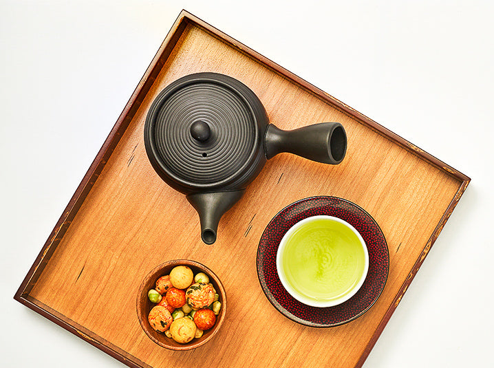 A cup of green tea in a white cup with black kyusu and Japanese snacks on a sakura wooden tray