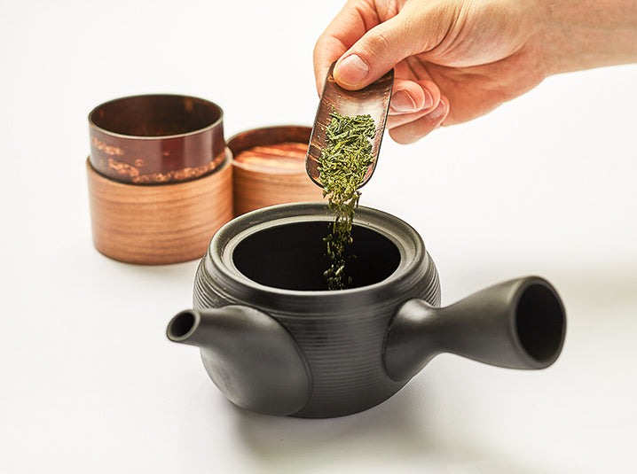 hand pouring green tea from sakura chasen teaspoon into a black kyusu teapot with sakura cherry blossom tea canisters in the background
