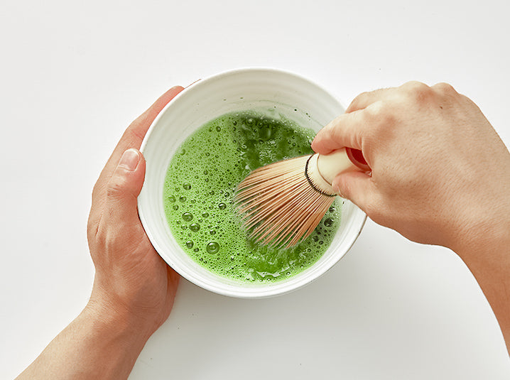 whisking ceremonial grade matcha powder in a white bowl using a bamboo whisk to make a cold dairy free oat milk matcha latte beverage