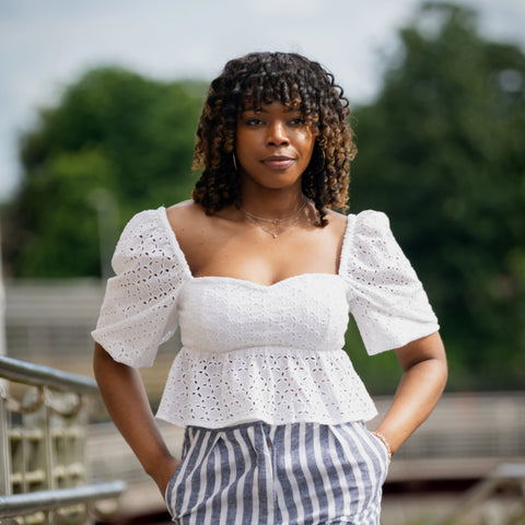 A woman with curly hair wearing the Karee Timeless Allure Awaits: White Lace Whisper Blouse and striped pants stands outdoors, hands in pockets, with a neutral expression. Trees and a railing are in the blurred background. Her outfit exudes a romantic charm, effortlessly blending into her sustainable wardrobe.