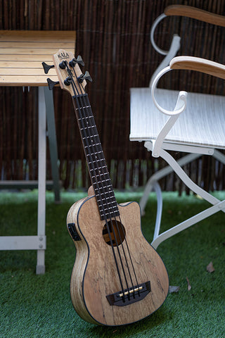 Kala U•BASS® Built with Spalte Maple Wood, leaning against a wood-top patio table with white metal legs on artificial grass; a white patio chair is in the background