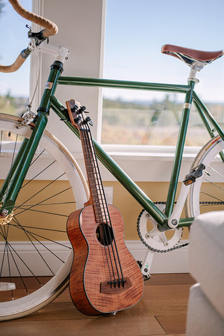 A Kala U•BASS® built from Exotic Mahogany wood, leaning agains a green bicycle and a brightly lit window.