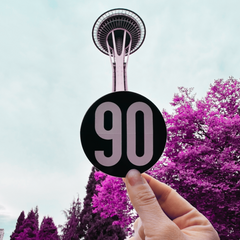 A sticker with the number 90 brand logo held up against the backdrop of Seattle's iconic Space Needle, symbolizing the brand's grunge heritage.