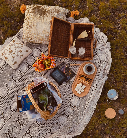 Aesthetic picnic setup with a lace blanket, a smores tray, picnic basket, snacks and cute pillows