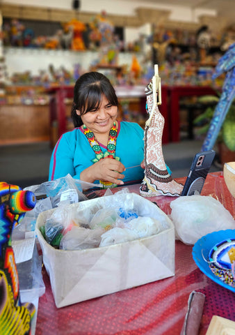 Una artesana huichol en un mercado de artesanías sonríe mientras trabaja en un objeto de arte decorativo. Luce una blusa turquesa vibrante y un collar de cuentas multicolores, complementando su atuendo tradicional. Sostiene una escultura detallada en proceso, con diseños intrincados de chaquira en blanco, negro y marrón. La mesa delante de ella está desordenada con materiales de arte, como cuentas y hilos. En el fondo borroso, se ven más coloridas artesanías huicholas, transmitiendo una atmósfera alegre y creativa