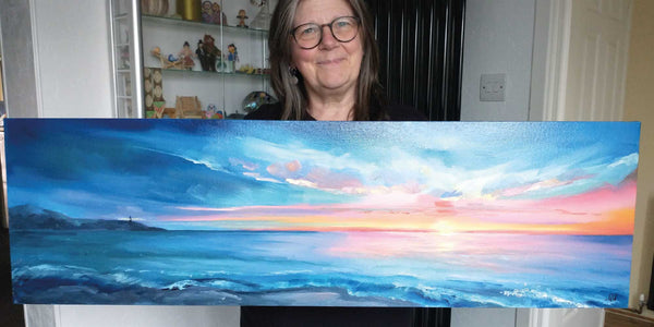 Picture of a woman holding a landscape oil painting of a beach and skyscape by Georgina Westley