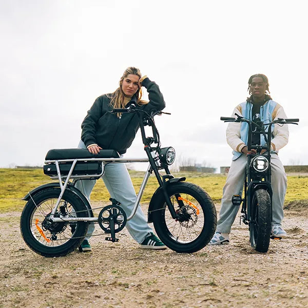 Two youths wearing urban clothing, one girl and one boy, posing with two electric fatbikes in the sand