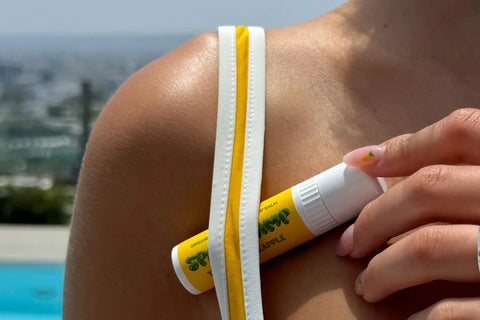 Tropical Pineapple Space Camp flavored lip balm held near a sunny poolside shoulder with yellow swimwear straps