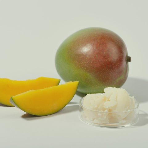 A vibrant, whole mango fruit next to a transparent glass bowl filled with medium-sized cubes of mango butter, and two upright slices of mango fruit playfully arranged like a smile.