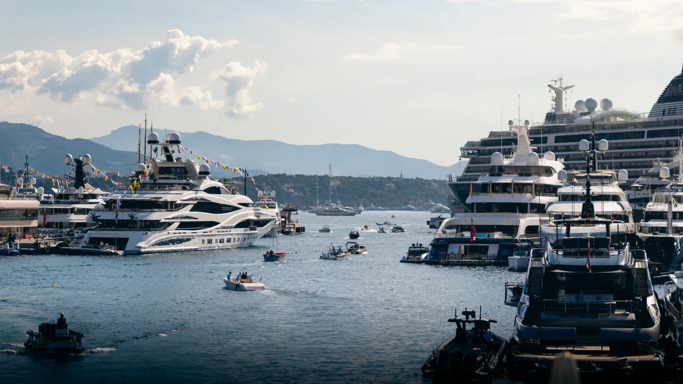 Luxury yachts docked in Monaco's Port Hercules during the F1 Grand Prix, with small boats on the water and mountains in the background