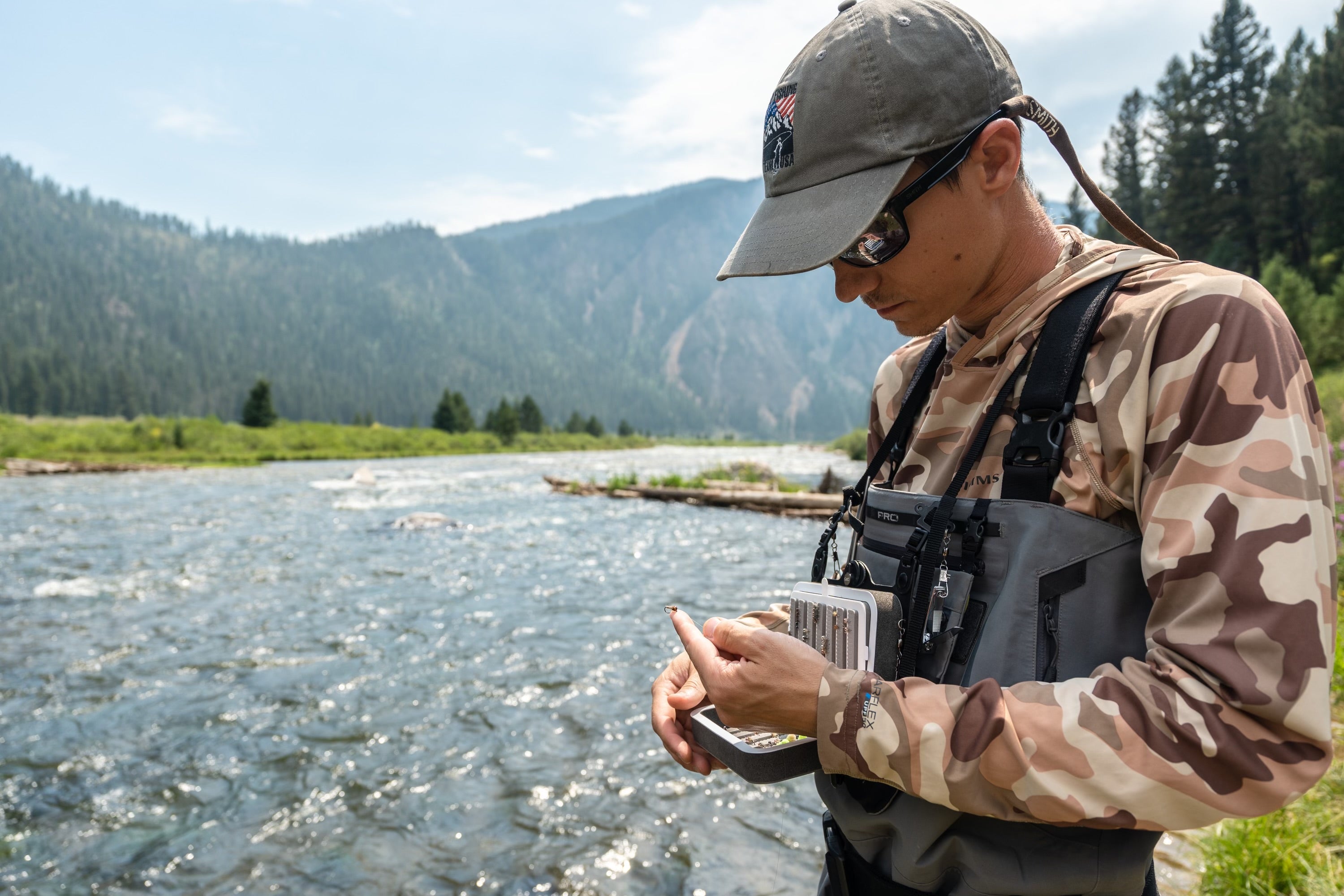 Josh Miller tying a fly to his line with a ball cap and waders on standing in a river with a mountain landscape back drop.