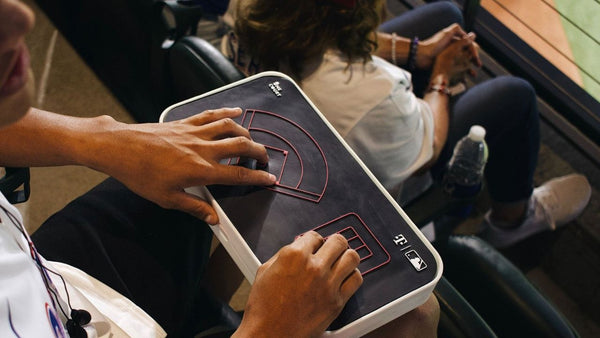 The image shows a person using OneCourt’s haptic mini-field device at a sports game. The device is a rectangular, tablet-like object designed to allow users, particularly those with visual impairments, to experience the layout of a baseball field through touch. It features raised, tactile outlines of a baseball diamond and other key field areas, marked with red lines for enhanced contrast. The person is sitting in a stadium seat, with their hands placed on the device, feeling the haptic field to follow the game's layout or events. In the background, another spectator is seated, watching the game. The device likely enhances the accessibility of the game experience for blind or visually impaired users.