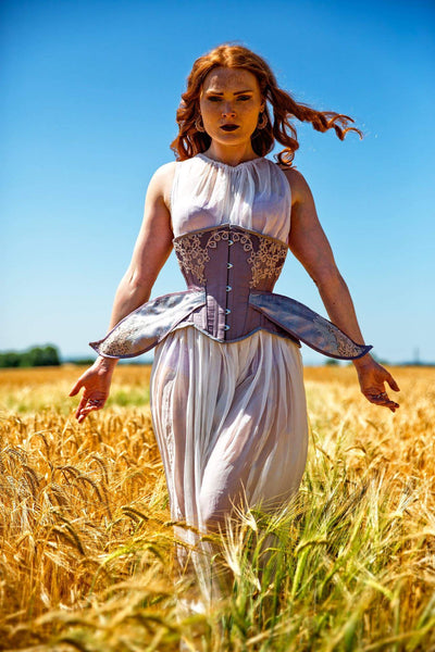 Model stands in a sunny field wearing a lilac corset with wings