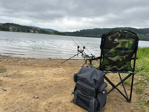 Angelzubehör am Seeufer mit einem Tarnstuhl und angelbereiten Angelruten, ruhiger See und bewölkter Himmel im Hintergrund