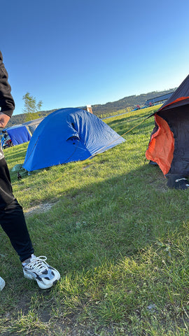 Campingplatz mit blauen und orangefarbenen Zelten auf einer Wiese am Seeufer an einem sonnigen Tag, malerische Hügel im Hintergrund.