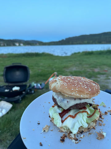 Selbstgemachter Burger mit Salat, Käse und Sauce auf einem Sesambrötchen, serviert auf einem blauen Teller mit unscharfem Campinghintergrund am Seeufer."