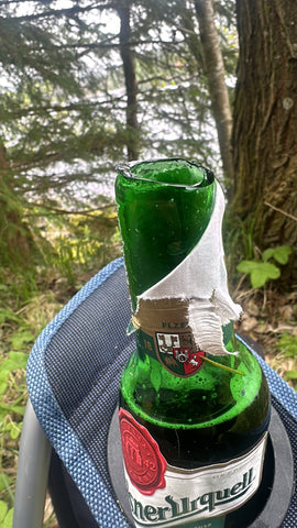 Nahaufnahme einer grünen Bierflasche mit weißem Etikett auf einem blauen Campingstuhl, umgeben von Bäumen in einer Waldumgebung.