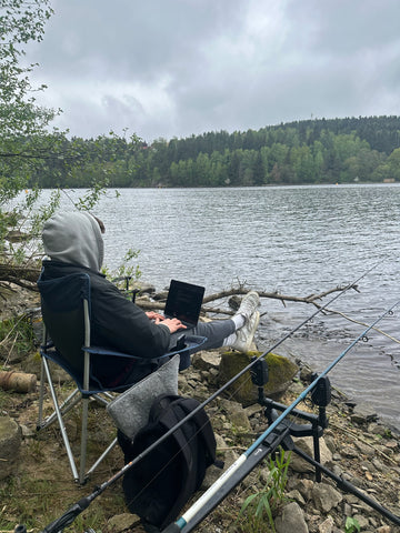 Mann sitzt auf einem Klappstuhl am Seeufer, arbeitet am Laptop und angelt gleichzeitig, Angelruten neben ihm aufgestellt, ruhiger waldreicher See im Hintergrund