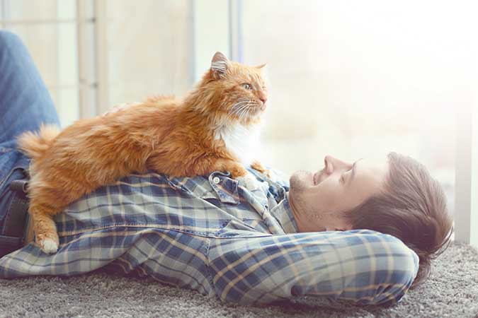 a young handsome man, laying on the floor on his back. He has a orange long-haired cat sitting on top of him. They are looking at each other