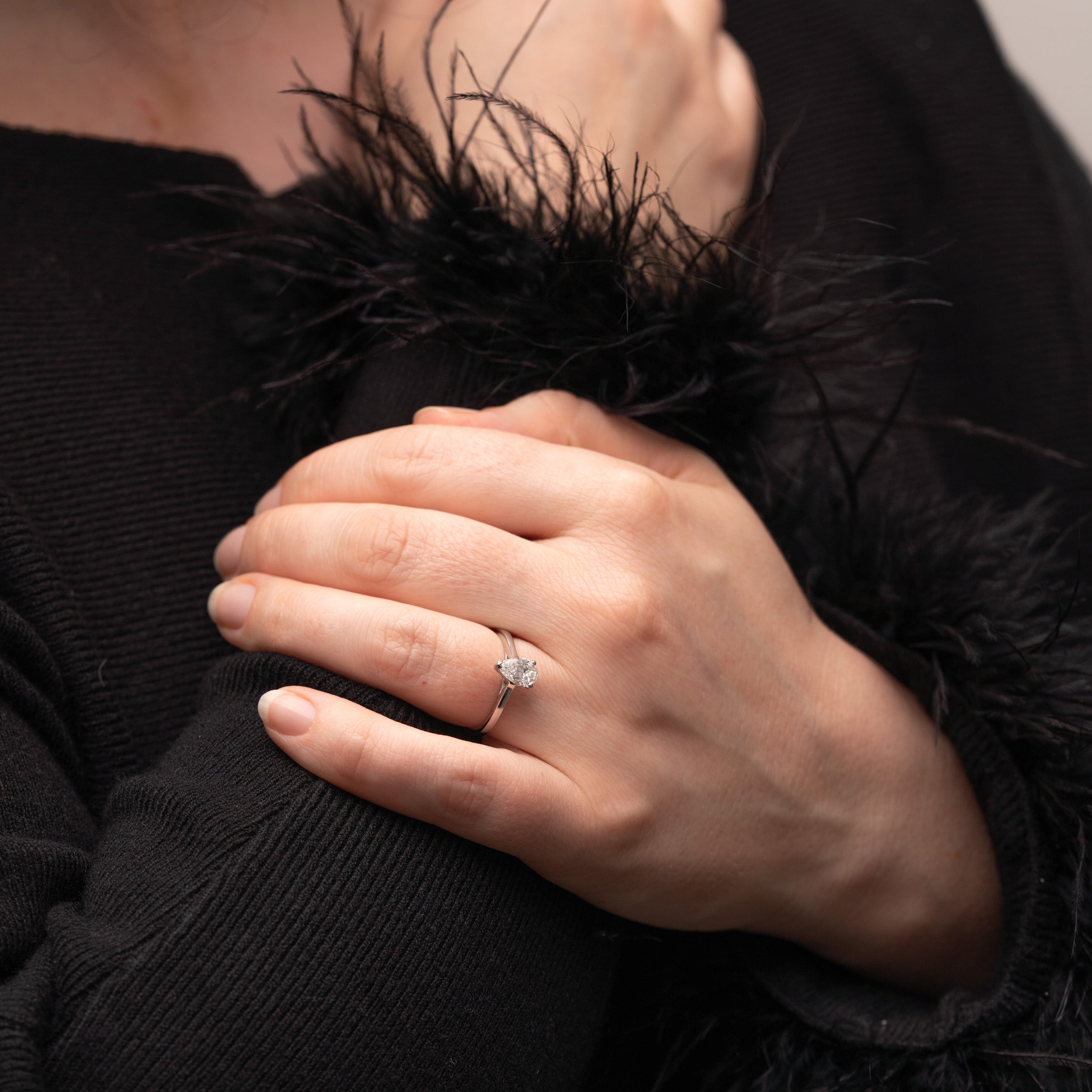 Slim band solitaire pear cut ring is worn on models hand, model is wearing black jumper with black feather sleeves.