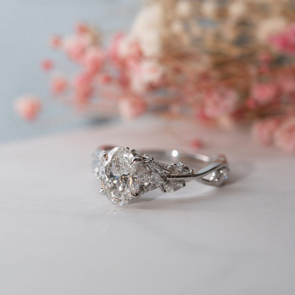 Image of multistone nature inspired engagement ring with oval centre stone set in Platinum facing front/side on a white and blue background with pink dried flowers.