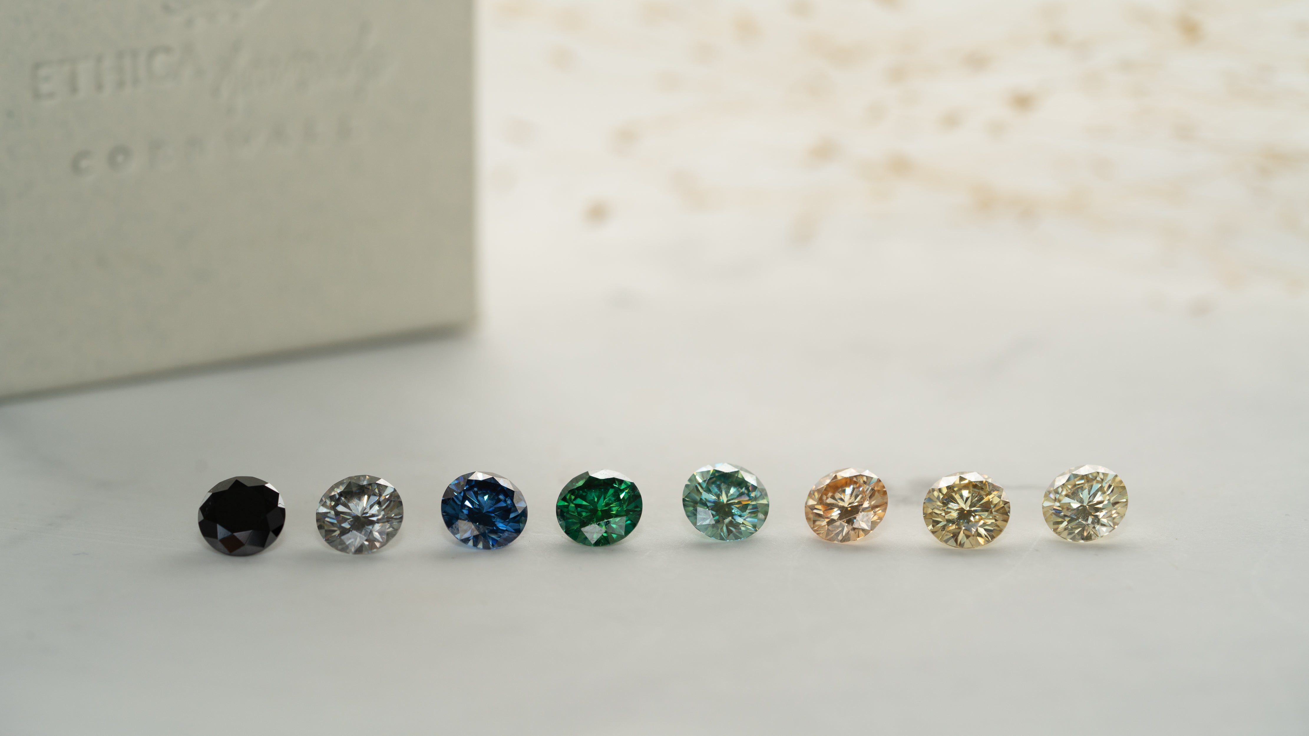 Eight Ethica Diamond moissanite loose stones in eight different colours are photographed in a line on a soft white background, with ring box and dried leaves in the background.
