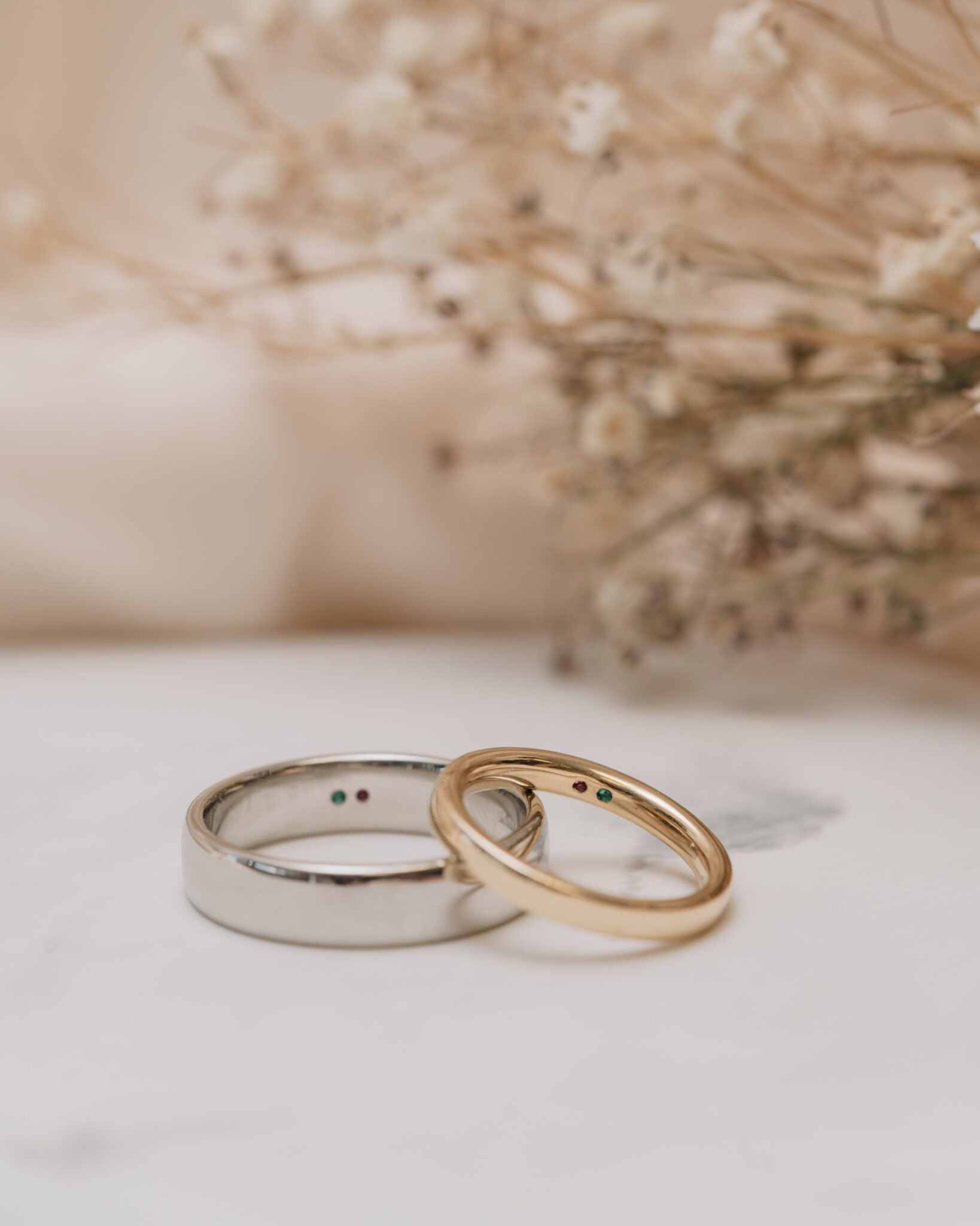 two wedding rings resting on a plain white surface with flowers in the background and small gemstones set on the inside of the bands