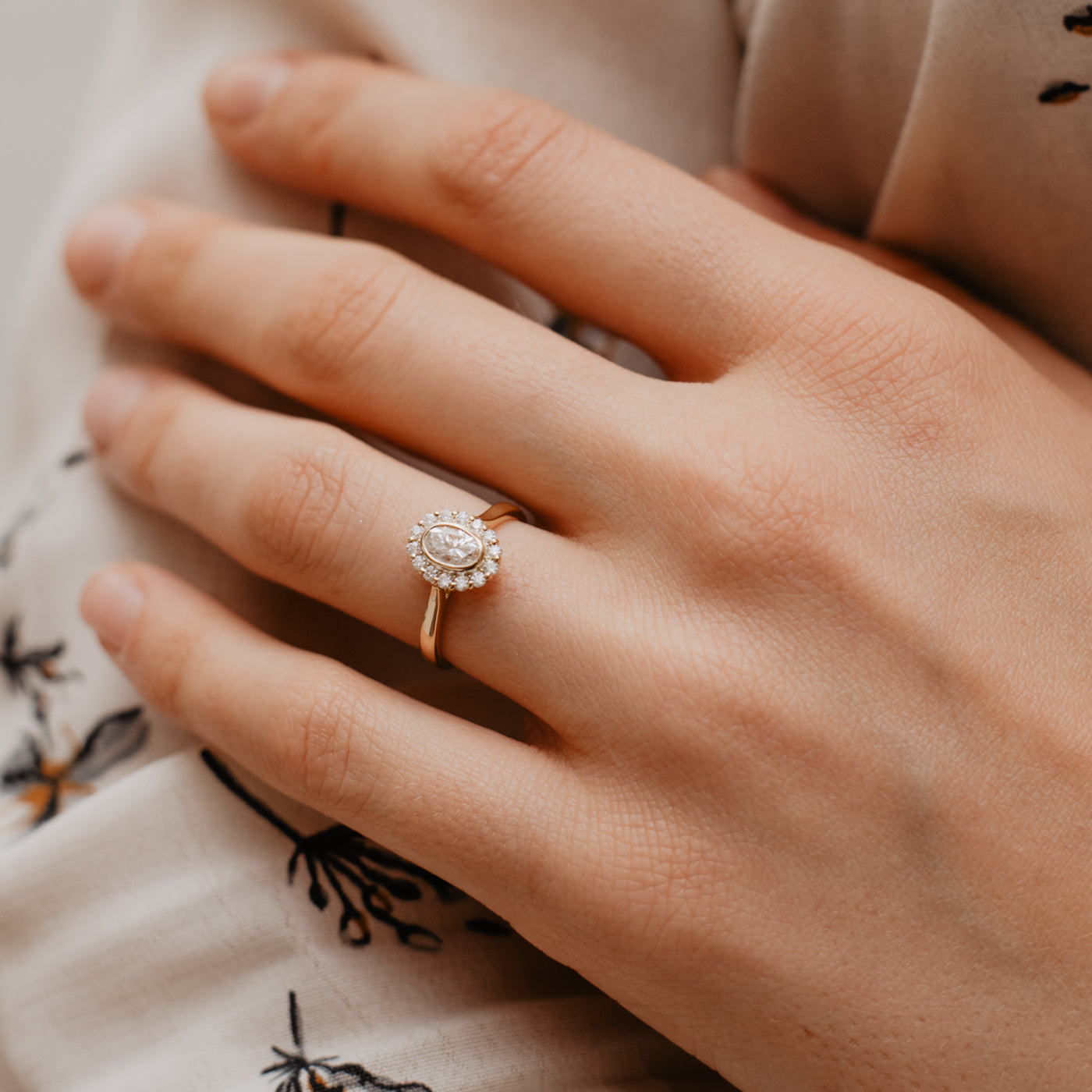 Lenore oval halo ring worn on models hand, her hand is resting on her arm and she's wearing a cream flowery blouse.