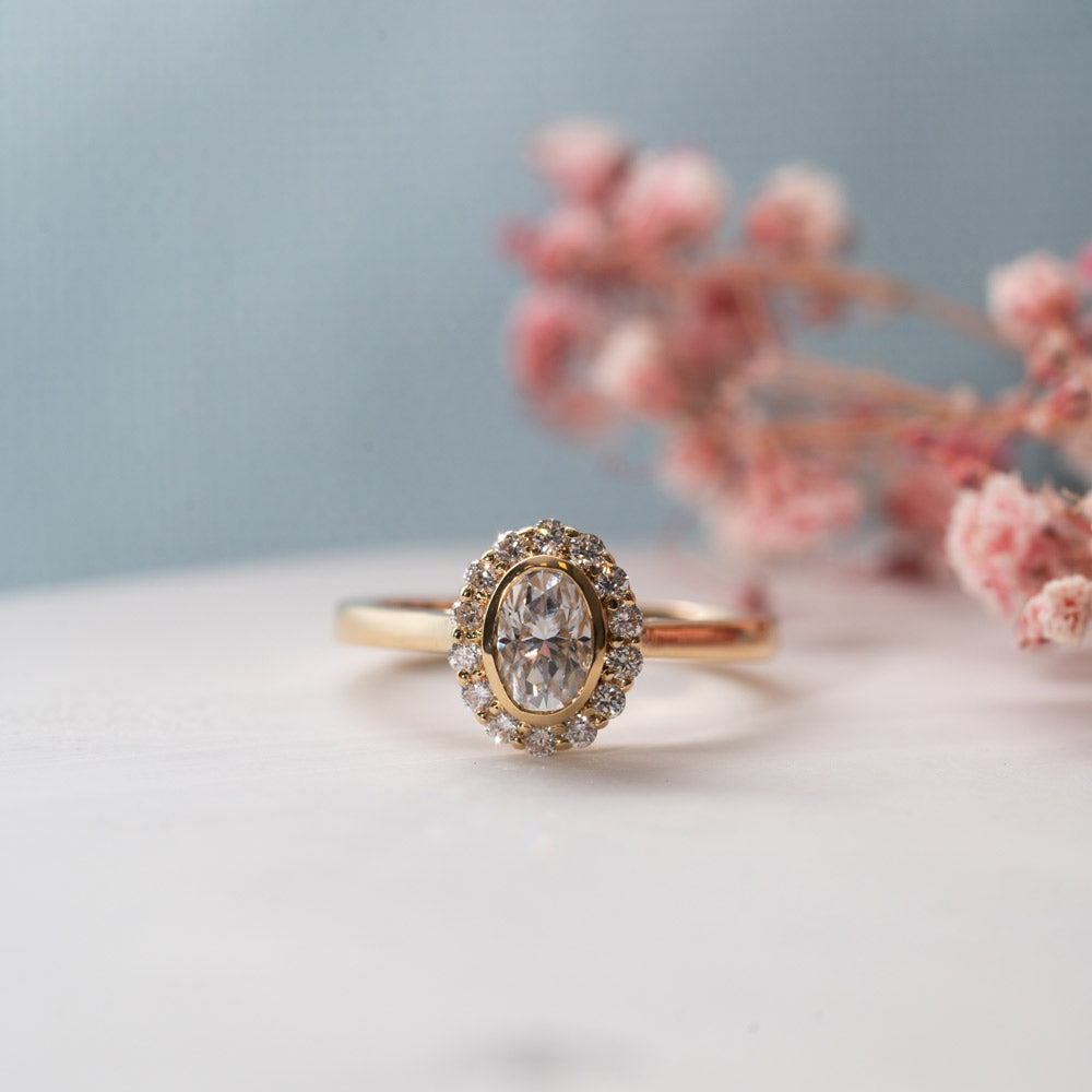 Oval halo engagement ring in yellow gold on a white and blue background.