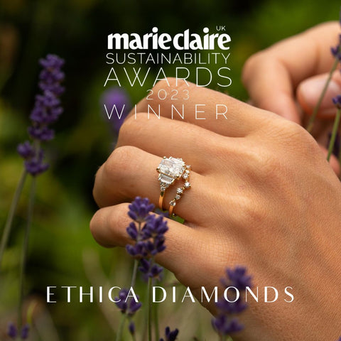 Photograph of accented emerald cut engagement ring with multi-stone stacking ring in gold worn on models hand surrounded by lavender. 