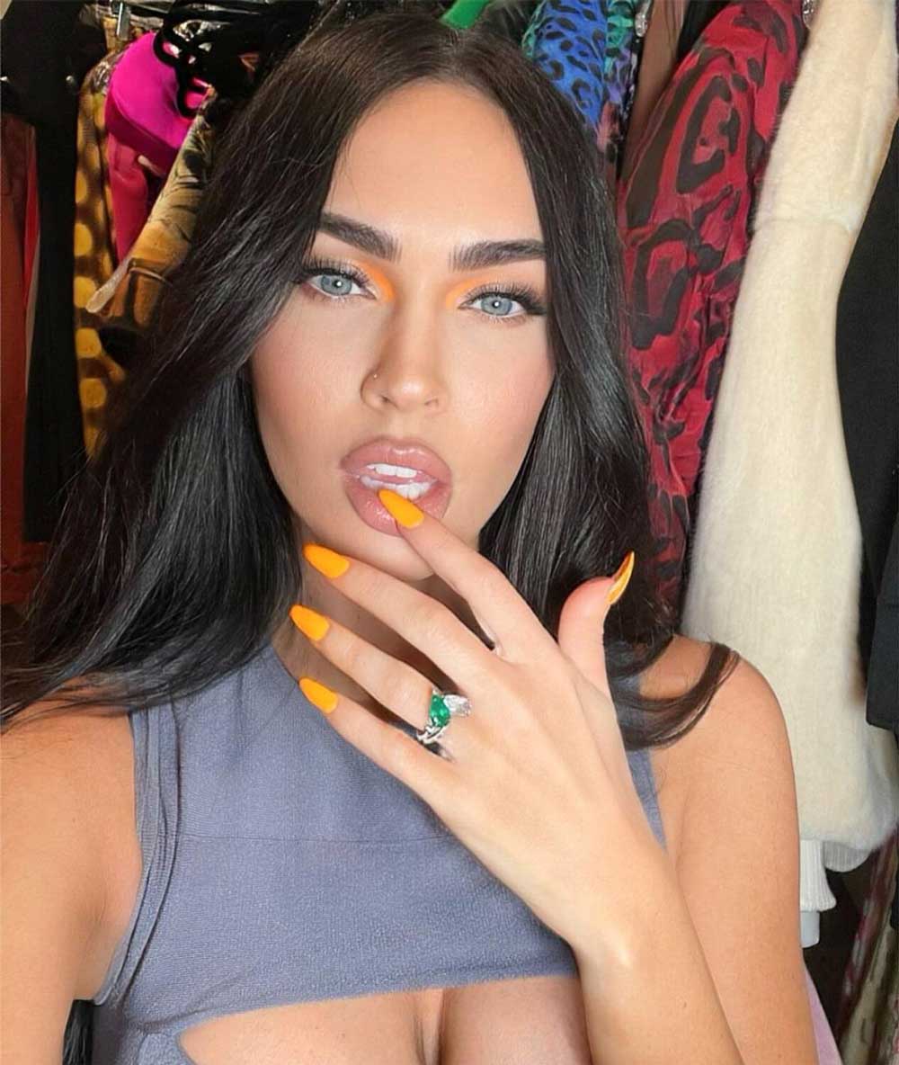 A selfie of Megan with her hand up by her face, showing off her long orange nails and engagement ring.