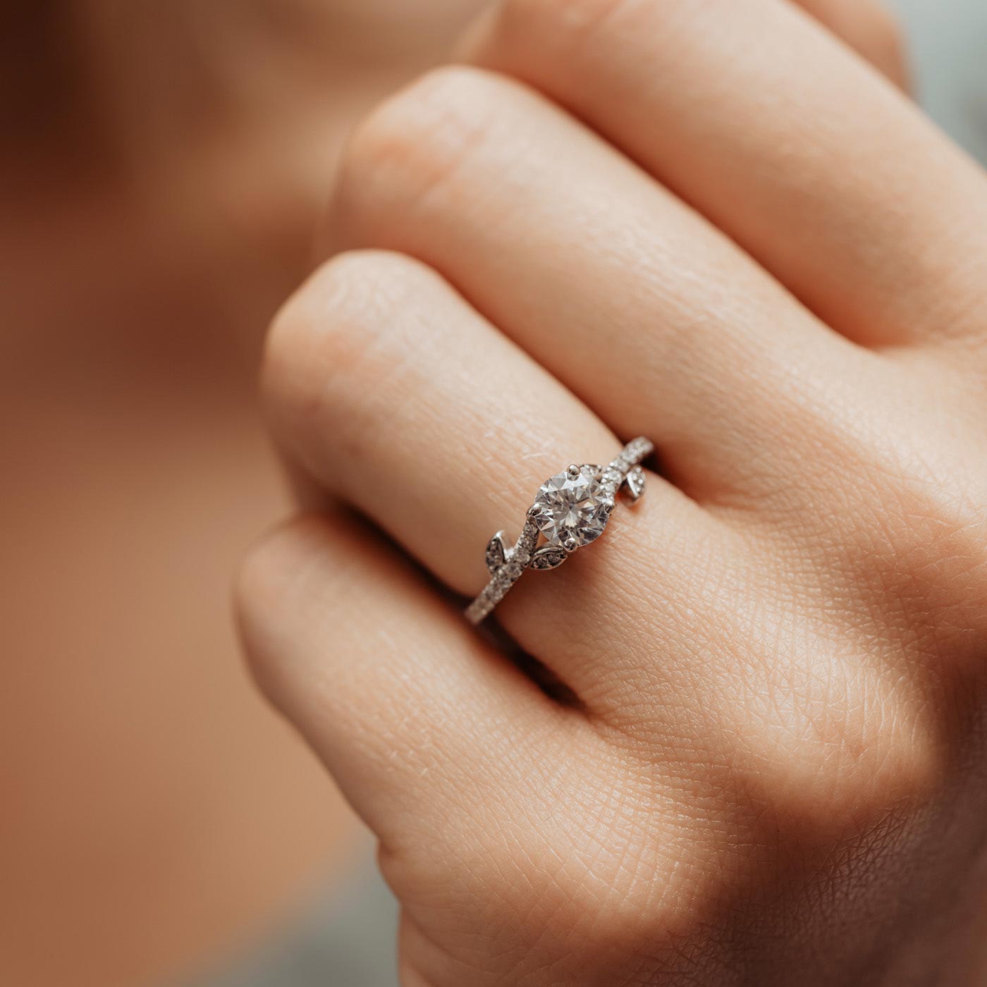 Close up of nature inspired engagement ring with round centre stone and stone set shoulders with leaf shaped accents worn on models hand.