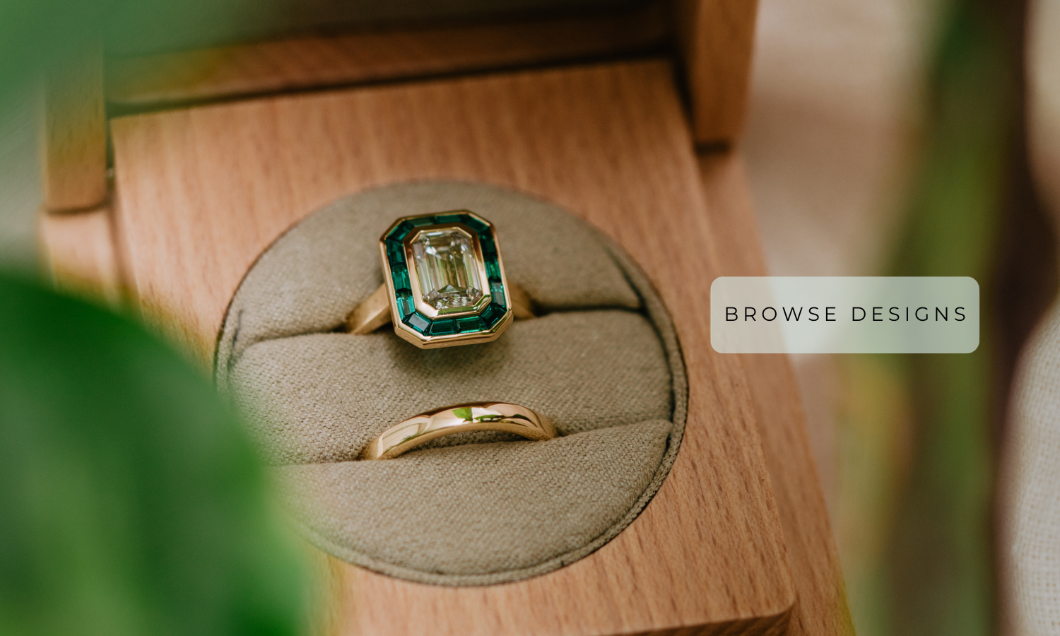 Close up photograph of an emerald cut halo engagement ring with green emerald halo and matching gold wedding band in a wooden box with green leaves. 