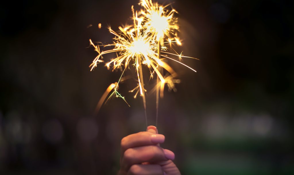 Image of a hand holding two lit sparklers up against a night background.