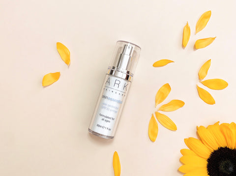 Product Image of ARK Skincare's SPF 30 Primer on a cream background next to a sunflower and vibrant yellow petals.