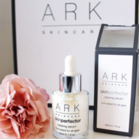 Product image of ARK Skincare's Clearing serum next to it's product box and a pretty pink rose, laid out in front of a branded ARK Skincare gift box.