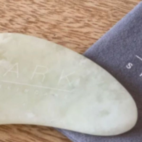 Product Image of ARK Skincare's Gua Sha Jade Tool laid on a wooden table with it's grey velvet drawstring bag