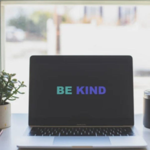 Image of a laptop screen with a 'be kind' screen saver, on a desk with coffee and a plant