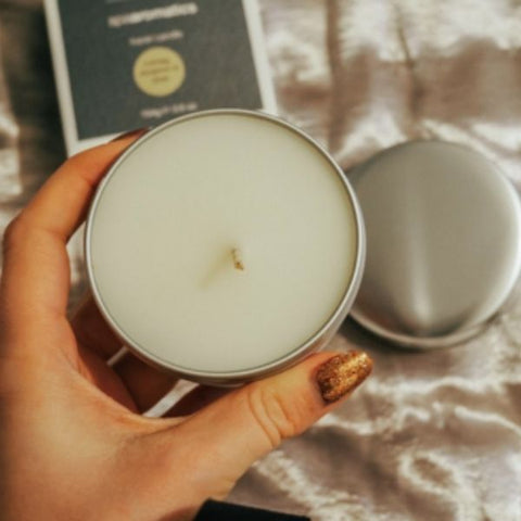 Product Image of ARK Skincare's Travel Candle being held above a cosy looking bed sheet with its branded product box.  
