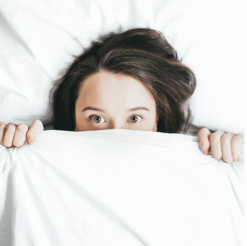 Image: Woman tucked up in bed behind white bed sheets held up to underneath her eyes