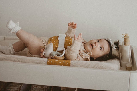 A baby with dark brown hair is lying on a change table, wrapped in a WriggleBum nappy change harness. The baby is kicking their legs.