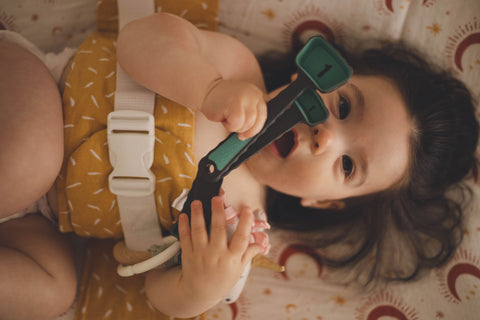 A baby with dark brown hair is lying down on a change table, wrapped in a WriggleBum nappy change harness. They are playing with a set of plastic measuring spoons.