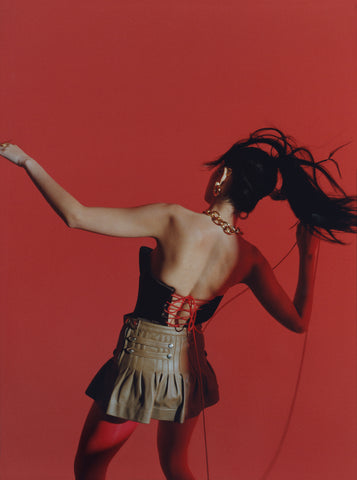 Woman facing away from the camera against red background, wearing gold chain necklace, red top, and skirt